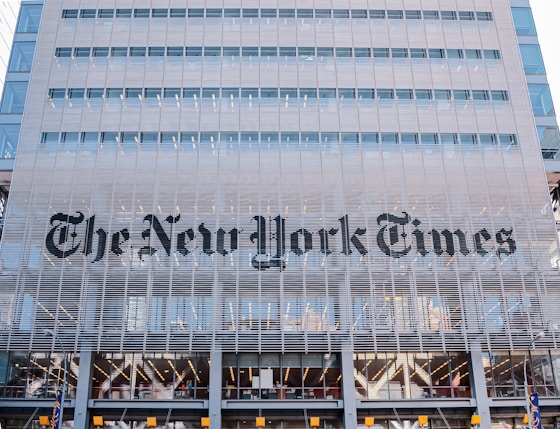 The building facade features large, modern architectural elements with a grid-like structure. The name of a well-known newspaper is prominently displayed in bold, black letters across the midsection. The structure consists of multiple floors with visible windows, suggesting it is an office building. The design incorporates a sleek and contemporary style.