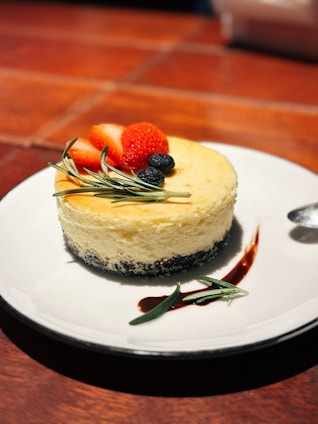 A cheesecake with a creamy texture, topped with fresh strawberries, blueberries, and sprigs of rosemary. It is served on a white plate with a dark, crumbly base. The setting includes a brown, tiled table surface, and the edge of a spoon is visible on the right side of the plate.