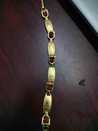 A bracelet with gold-colored segments connected by small U-shaped links. Each segment features intricate designs or characters etched into the surface, and the links contain an orangish-brown stone. The bracelet is laid out on a dark wooden surface next to a screw or small bolt.