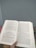 A person holds an open book with text written in a language resembling Hindi or Sanskrit. The book is held in a well-lit environment with a neutral wall in the background and a glimpse of a plant with small, pink flowers.