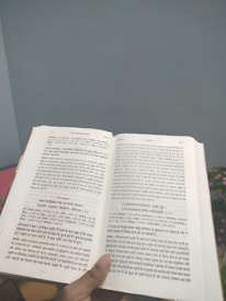 A person holds an open book with text written in a language resembling Hindi or Sanskrit. The book is held in a well-lit environment with a neutral wall in the background and a glimpse of a plant with small, pink flowers.