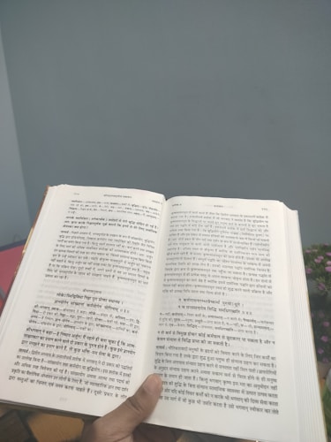 A person holds an open book with text written in a language resembling Hindi or Sanskrit. The book is held in a well-lit environment with a neutral wall in the background and a glimpse of a plant with small, pink flowers.