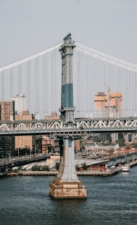 A large suspension bridge spans across a river, with urban buildings in the background. The bridge features a prominent steel tower with intricate design elements. Cars are visible on the roadway of the bridge. The cityscape includes high-rise buildings and construction sites with cranes.