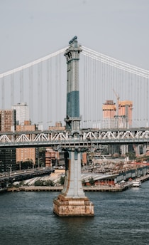A large suspension bridge spans across a river, with urban buildings in the background. The bridge features a prominent steel tower with intricate design elements. Cars are visible on the roadway of the bridge. The cityscape includes high-rise buildings and construction sites with cranes.