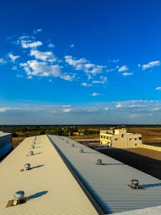 A large industrial rooftop with ventilation turbines is seen extending towards the horizon under a bright blue sky scattered with fluffy white clouds. In the background, there is a flat landscape with sparse vegetation and a distant building, possibly an industrial facility.