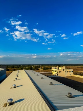 A large industrial rooftop with ventilation turbines is seen extending towards the horizon under a bright blue sky scattered with fluffy white clouds. In the background, there is a flat landscape with sparse vegetation and a distant building, possibly an industrial facility.
