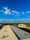 A large industrial rooftop with ventilation turbines is seen extending towards the horizon under a bright blue sky scattered with fluffy white clouds. In the background, there is a flat landscape with sparse vegetation and a distant building, possibly an industrial facility.