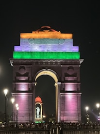 A monumental arch structure brightly illuminated with vibrant tricolor lighting. The top of the arch displays orange, white, and green lights, reminiscent of the Indian flag. Surrounding street lamps cast a gentle glow and a large sculpture is visible through the central arch. The scene is bustling with people and activity in the foreground.