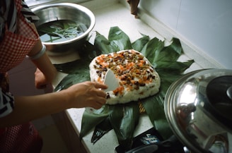 A person is preparing a dish on a kitchen countertop. The dish consists of a spread of leafy greens topped with a mixture of rice, nuts, and dried fruits. A spatula is being used to serve or arrange the ingredients. There is a metal pot with a lid on the right and a bowl with water and leaves to the side.