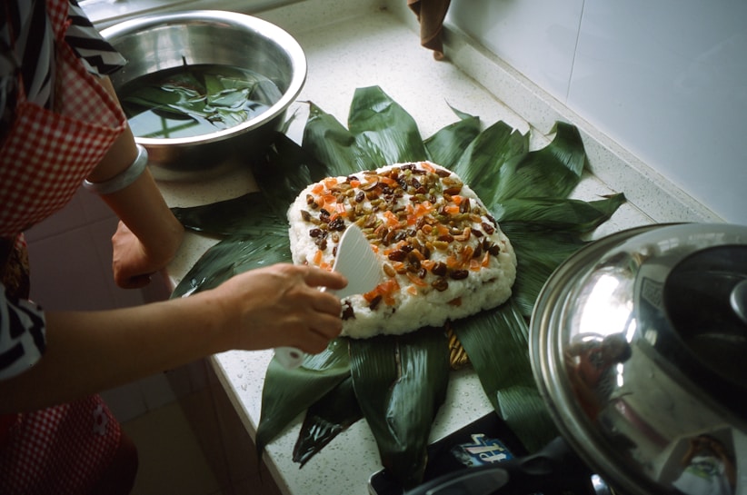A person is preparing a dish on a kitchen countertop. The dish consists of a spread of leafy greens topped with a mixture of rice, nuts, and dried fruits. A spatula is being used to serve or arrange the ingredients. There is a metal pot with a lid on the right and a bowl with water and leaves to the side.