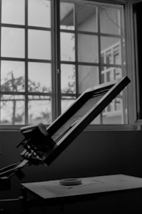 A drafting table with a propped-up drawing board is in the foreground, positioned next to a large window with grid-like panes. Natural light streams in through the window, casting soft shadows on the table and emphasizing the indoor setting.
