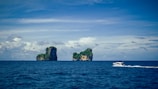 Over a vast expanse of deep blue ocean, two small rocky islands covered in lush green vegetation rise prominently. The sky above is partly cloudy with varying shades of blue and white. A speedboat zips across the water in the foreground, leaving a trail of white waves in its wake.