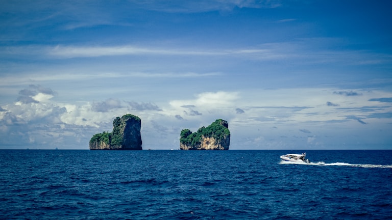 Over a vast expanse of deep blue ocean, two small rocky islands covered in lush green vegetation rise prominently. The sky above is partly cloudy with varying shades of blue and white. A speedboat zips across the water in the foreground, leaving a trail of white waves in its wake.
