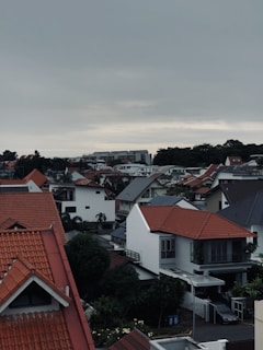 A neighborhood with a multitude of houses featuring mostly red-tiled roofs. The buildings are mostly multi-story homes with modern architectural design elements. It is an overcast day with thick, gray clouds covering the entire sky. Trees and greenery are scattered throughout the area, giving it a suburban feel.