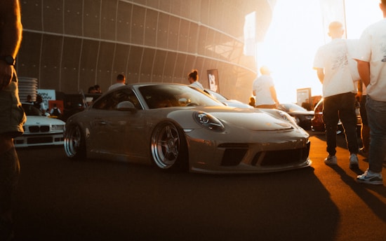 A sleek sports car with a glossy finish is parked among a group of people at what appears to be a car show or gathering. The scene is warmly lit by the setting sun, casting long shadows on the ground. Several people are standing around, casually dressed, and engaged in conversation.