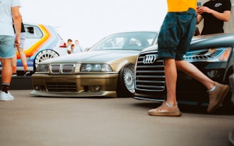 A group of people are gathered around and admiring several cars, including a gold BMW and a black Audi prominently displayed. There is also a colorful, rainbow-painted vehicle in the background. The scene likely takes place at a car show, with people casually dressed, some taking pictures and others simply observing the cars.