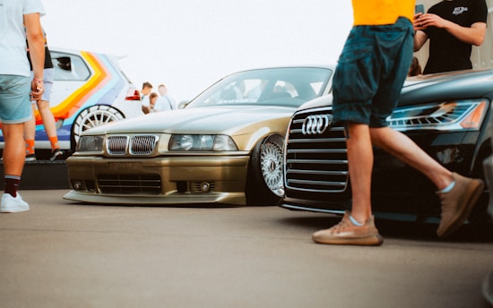A group of people are gathered around and admiring several cars, including a gold BMW and a black Audi prominently displayed. There is also a colorful, rainbow-painted vehicle in the background. The scene likely takes place at a car show, with people casually dressed, some taking pictures and others simply observing the cars.