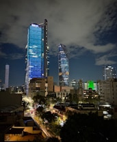 A night-time cityscape featuring two prominent skyscrapers with one notably illuminated in blue and marked with the logo 'BBVA'. The skyline includes other modern buildings with various lights and colors, along with a busy street below filled with traffic and trees lining the road. The sky is partly cloudy, adding a dramatic backdrop to the scene.