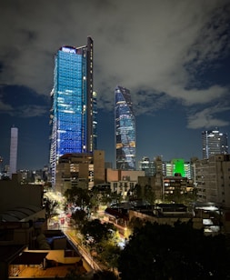 A night-time cityscape featuring two prominent skyscrapers with one notably illuminated in blue and marked with the logo 'BBVA'. The skyline includes other modern buildings with various lights and colors, along with a busy street below filled with traffic and trees lining the road. The sky is partly cloudy, adding a dramatic backdrop to the scene.