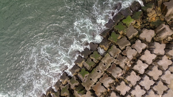 Aerial view of ocean waves crashing against large, star-shaped concrete tetrapods on a coastline. Some of the tetrapods are covered in green moss, highlighting the interaction between man-made structures and natural elements.