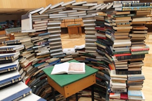A large number of books are stacked to form an arch-like structure, creating a small central nook with a table. An open book is placed on the table which is covered with a green felt surface. The surrounding area appears to be in a library, with more books visible in the background, along with card catalog drawers.