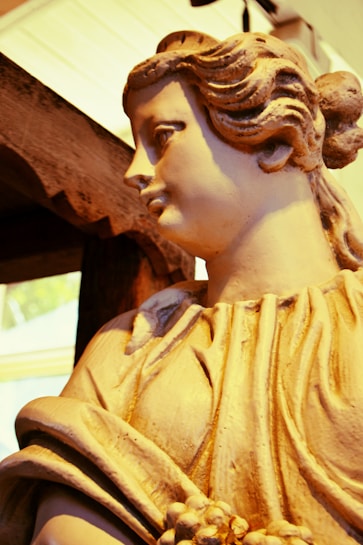 A detailed statue of a classical figure made of stone or plaster. The figure has wavy hair pulled back and wears a draped garment. The surface appears to have a warm, beige tone with visible texture. A partially visible wooden or rustic frame is positioned nearby.