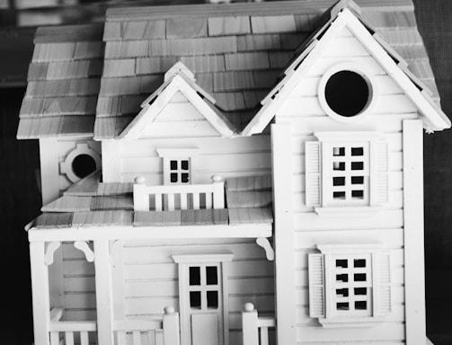A detailed scale model of a house, featuring intricate architectural elements. The miniature structure includes a gabled roof, window shutters, and a small balcony or porch area. The craftsmanship suggests a classic, possibly Victorian style.