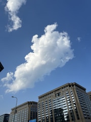 Large, modern buildings with reflective glass windows stand under a clear blue sky. A prominent, fluffy white cloud drifts above, casting subtle reflections on the buildings below.