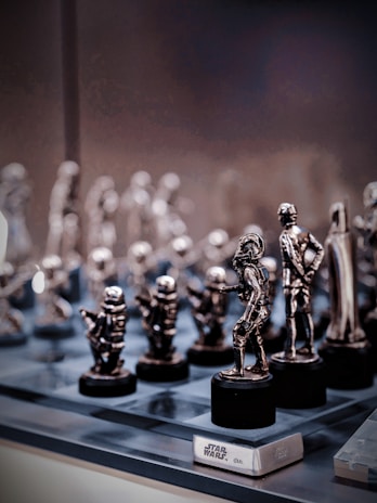 A collection of small, metallic figurines displayed on a reflective surface. These figurines resemble characters from the Star Wars universe, intricately crafted and placed on black pedestals. The background is softly blurred, emphasizing the shiny and detailed figures in the foreground.