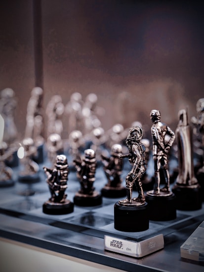A collection of small, metallic figurines displayed on a reflective surface. These figurines resemble characters from the Star Wars universe, intricately crafted and placed on black pedestals. The background is softly blurred, emphasizing the shiny and detailed figures in the foreground.