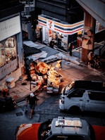 A street scene in an urban environment with a busy convenience store on the corner. Outside, there are several stalls displaying various goods under warm lighting. Vehicles, including a taxi and a van, are on the road, and pedestrians are moving about, adding to the city's bustling atmosphere.