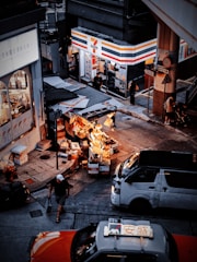 A street scene in an urban environment with a busy convenience store on the corner. Outside, there are several stalls displaying various goods under warm lighting. Vehicles, including a taxi and a van, are on the road, and pedestrians are moving about, adding to the city's bustling atmosphere.