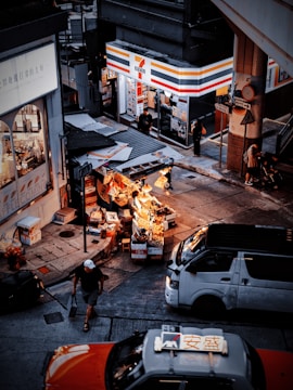 A street scene in an urban environment with a busy convenience store on the corner. Outside, there are several stalls displaying various goods under warm lighting. Vehicles, including a taxi and a van, are on the road, and pedestrians are moving about, adding to the city's bustling atmosphere.