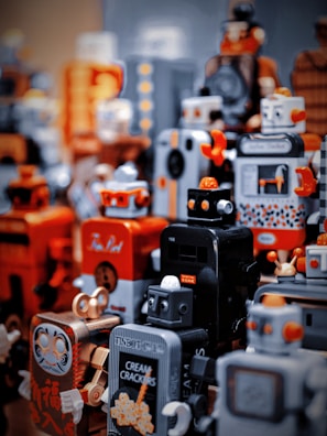 A collection of toy robots with a retro aesthetic. The robots are predominantly orange, black, and gray, featuring various designs and facial expressions. Some have intricate patterns and detailing resembling vintage machinery.