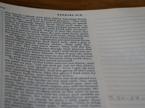 A close-up view of a page from a Bible open to the book of Ezekiel, with text visible and partially focused on verses discussing divine warnings and moral guidance. The page appears to be on a desk, and part of the page is slightly out of focus.