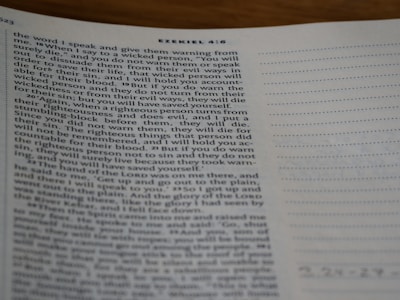 A close-up view of a page from a Bible open to the book of Ezekiel, with text visible and partially focused on verses discussing divine warnings and moral guidance. The page appears to be on a desk, and part of the page is slightly out of focus.
