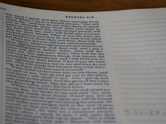 A close-up view of a page from a Bible open to the book of Ezekiel, with text visible and partially focused on verses discussing divine warnings and moral guidance. The page appears to be on a desk, and part of the page is slightly out of focus.