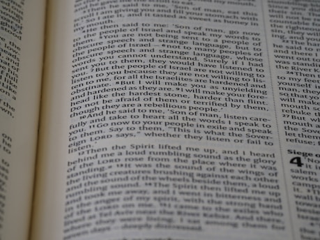 A close-up view of a page from a book, featuring text printed in a column layout. The text appears to be from a religious or philosophical work, with words and phrases presented in a structured format. The focus is on the middle of the page, with other text slightly blurred in the foreground and background.