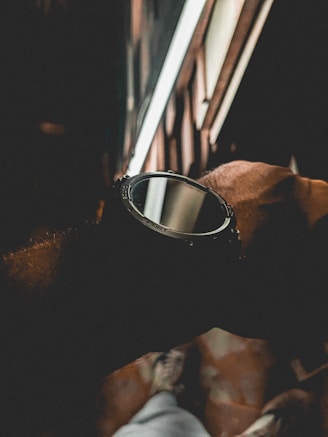 A close-up of a wristwatch with a sporty design. The watch is being worn on a wrist against a dimly lit backdrop featuring blurred patterns and lines. The lighting is subdued, creating a moody atmosphere.