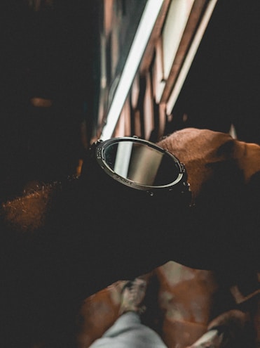 A close-up of a wristwatch with a sporty design. The watch is being worn on a wrist against a dimly lit backdrop featuring blurred patterns and lines. The lighting is subdued, creating a moody atmosphere.