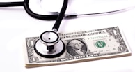 A stethoscope rests on top of a stack of one-dollar bills, suggesting a connection between healthcare and financial costs.