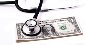 A stethoscope rests on top of a stack of one-dollar bills, suggesting a connection between healthcare and financial costs.