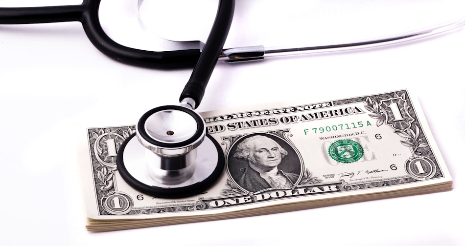A stethoscope rests on top of a stack of one-dollar bills, suggesting a connection between healthcare and financial costs.