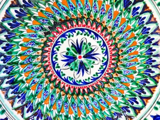 A vibrant and intricate circular pattern featuring layers of geometric and floral designs. The design predominantly uses blue, green, orange, and white colors, with detailed brush strokes and symmetrical shapes creating a mesmerizing mandala-like effect.