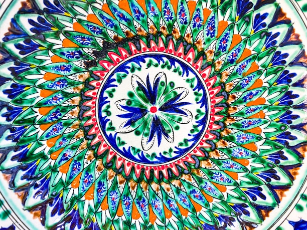 A vibrant and intricate circular pattern featuring layers of geometric and floral designs. The design predominantly uses blue, green, orange, and white colors, with detailed brush strokes and symmetrical shapes creating a mesmerizing mandala-like effect.