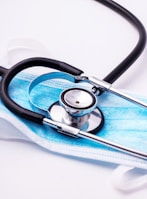 A stethoscope with black tubing is resting on top of a blue disposable face mask. The mask is set on a white surface, and the metallic part of the stethoscope reflects the surrounding light.