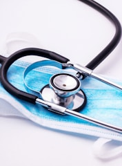 A stethoscope with black tubing is resting on top of a blue disposable face mask. The mask is set on a white surface, and the metallic part of the stethoscope reflects the surrounding light.