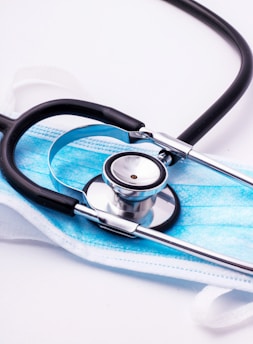 A stethoscope with black tubing is resting on top of a blue disposable face mask. The mask is set on a white surface, and the metallic part of the stethoscope reflects the surrounding light.