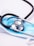A stethoscope with black tubing is resting on top of a blue disposable face mask. The mask is set on a white surface, and the metallic part of the stethoscope reflects the surrounding light.