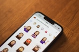 A smartphone displaying various Memoji stickers on its screen, placed on a wooden surface. The time and network details are visible at the top of the phone's screen.